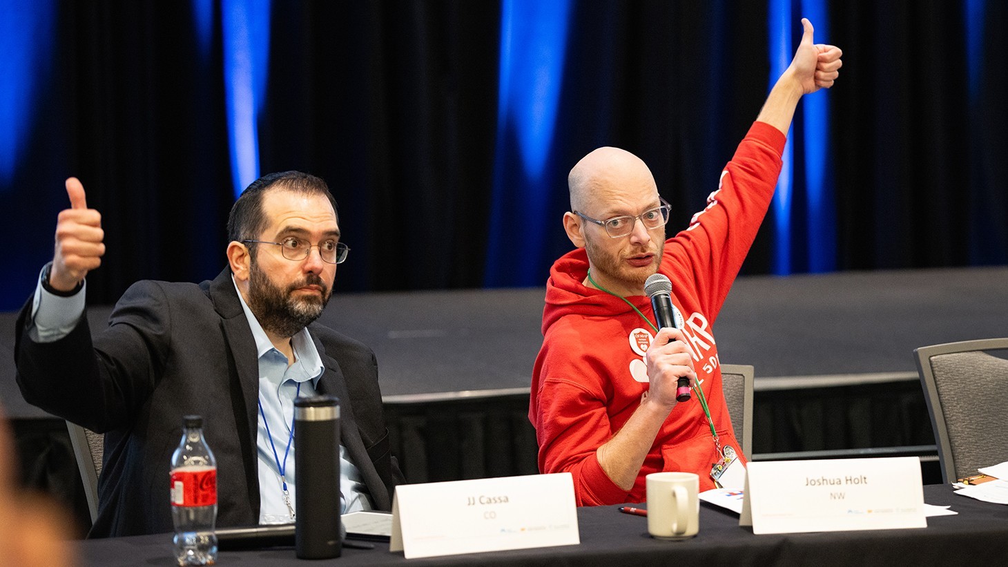 one man in a suit on left holds his thumb up and a man on the right wearing a red hooding holds his thumb up