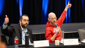 one man in a suit on left holds his thumb up and a man on the right wearing a red hooding holds his thumb up